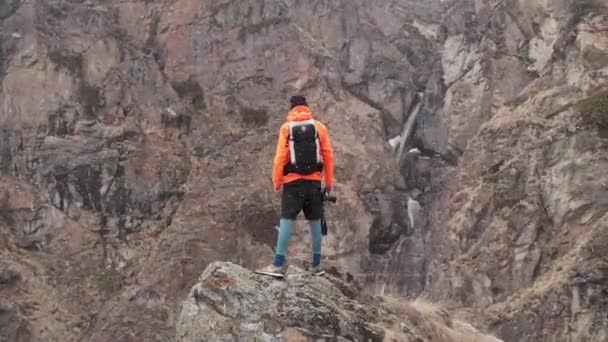 峡谷の高い岩の上にカメラを手にした若い男性写真家の空中写真と、春の吹雪と雪の中の高山の滝. — ストック動画