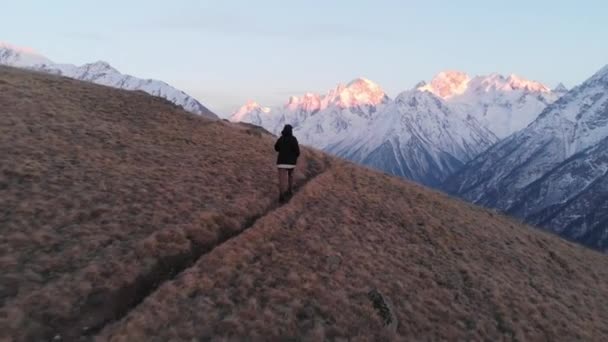 Een luchtfoto van een jong meisje in een pet loopt van een berg heuvel af naar de besneeuwde bergen na zonsondergang in het blauwe uur. Het concept van vreugde in de bergen van wandelen en joggen — Stockvideo