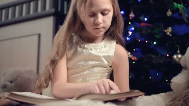 Ein kleines blondes Mädchen im festlichen Kleid mit einem Buch in der Hand sitzt neben Stofftieren vor dem Hintergrund eines Weihnachtsbaums und liest ein Buch, das die Seite mit ihrem Platz anführt.. — Stockvideo