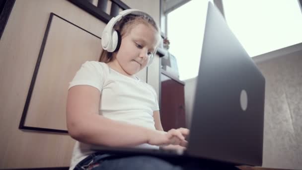 Una niña inteligente de siete años con auriculares blancos con un portátil en las manos está empujando al suelo en su habitación. La generación joven en Internet y la tecnología informática — Vídeo de stock