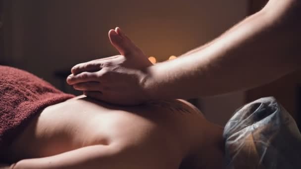 Close-up Jonge mannelijke massage therapeut doet rug massage aan een vrouw met een tatoeage in een massageruimte met schemerige lichten op de achtergrond van kaarsen. Low-key premium massageconcept — Stockvideo