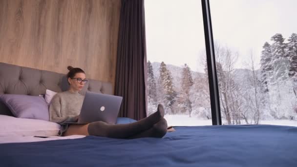 Retrato de atractiva chica freelancer en gafas con un portátil en la cama en un eco-hotel con paredes transparentes detrás de las cuales hay un bosque de invierno. Concepto de viaje y trabajo con horario libre. — Vídeos de Stock