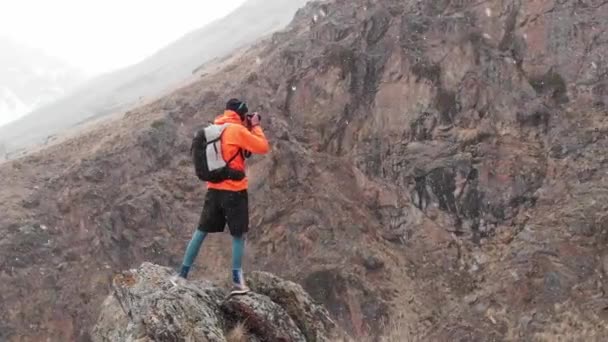 손에 카메라를 든 젊은 남자 사진 작가가 협곡에 있는 높은 바위 위에서 있는 모습과 봄에 눈보라와 눈 올 때 알프스 폭포 사진을 찍는 모습을 공중에서 목격 할 수있다. — 비디오