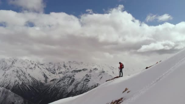 Vista aérea joven en gafas de sol con una mochila baja la montaña en la cresta en el borde de la nieve y las piedras contra el telón de fondo de las montañas cubiertas de nieve. — Vídeo de stock