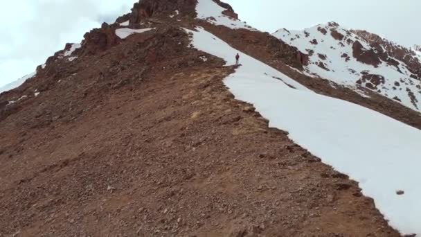 Een jonge man met een rugzak staat hoog in de bergen op de grens van sneeuw en rotsen op de kam tegen de achtergrond van besneeuwde bergen. Luchtfoto van het Alpengebied — Stockvideo