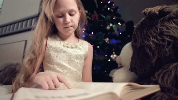 Mała blondynka w świątecznej sukience z książką w rękach siedzi obok miękkich zabawek na tle choinki i czyta książkę prowadzącą stronę z jej mieszkania. — Wideo stockowe