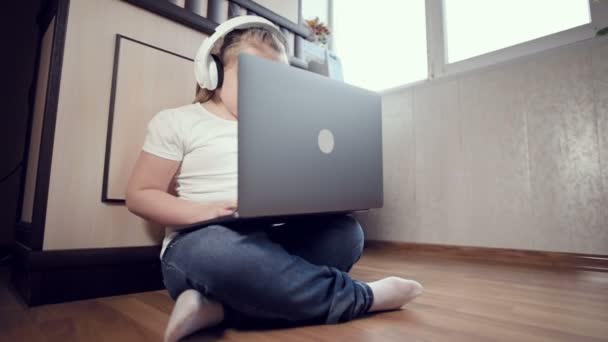 Ein schlaues kleines Mädchen von sieben Jahren mit weißen Kopfhörern und einem Laptop in der Hand schiebt sich in ihrem Zimmer auf den Boden. Die junge Generation im Internet und der IT-Technologie — Stockvideo