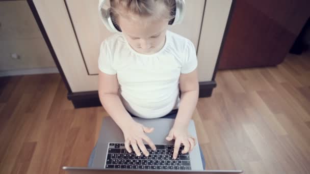 Розумна маленька семирічна дівчинка в білих навушниках з ноутбуком в руках штовхає підлогу в своїй кімнаті. Молоде покоління в Інтернеті і ІТ-технології — стокове відео