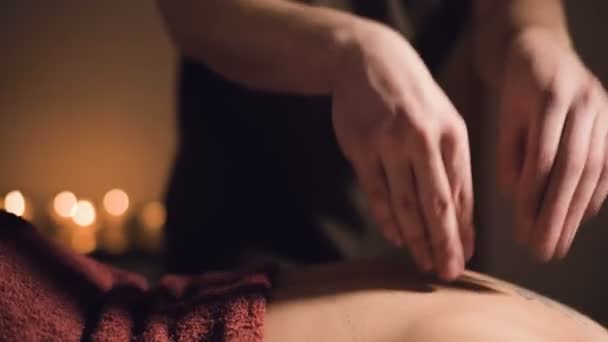 Nuori miespuolinen hieroja tekee naisen sormihierontaa takaisin tatuoinnilla hierontahuoneessa, jossa on hämärä valo kynttilöiden taustalla. Matala avain palkkio hieronta käsite — kuvapankkivideo