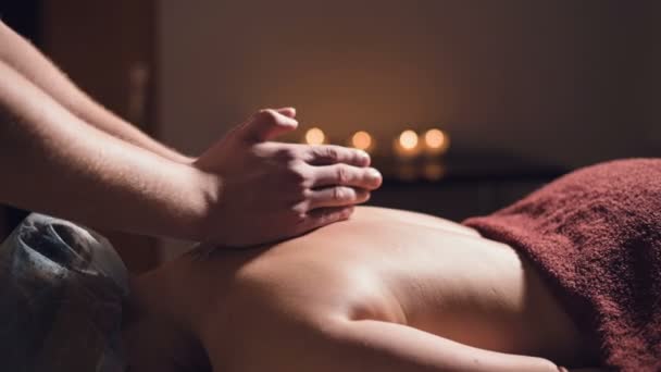 Close-up Jonge mannelijke massage therapeut doet rug massage aan een vrouw met een tatoeage in een massageruimte met schemerige lichten op de achtergrond van kaarsen. Low-key premium massageconcept — Stockvideo