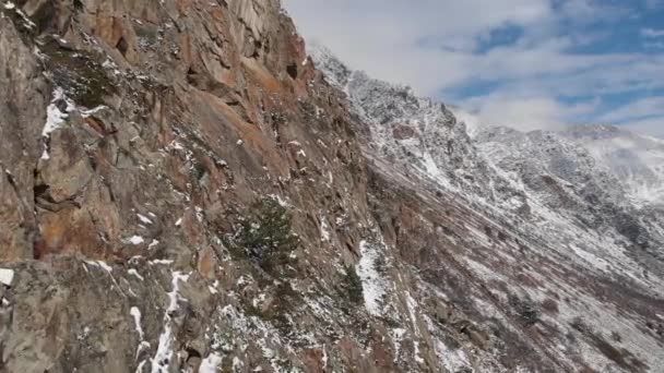 Αεροφωτογραφία που πετά δίπλα σε απότομους βράχους καλυμμένους με χιόνι σε ένα ορεινό φαράγγι μια ηλιόλουστη μέρα στις αρχές της άνοιξης. Η επική παράλλαξη στα βουνά του Βόρειου Καυκάσου — Αρχείο Βίντεο