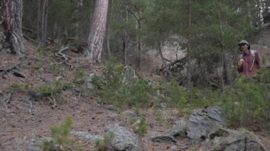 Sırt çantalı, uzun saçlı genç bir beyaz adam dağlardaki kozalaklı bir ormanda orman yolunda yürüyor.