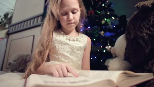 Een klein blond meisje in een feestelijke jurk met een boek in haar handen zit naast zacht speelgoed tegen de achtergrond van een kerstboom en leest een boek leidt de pagina met haar plaats. — Stockvideo