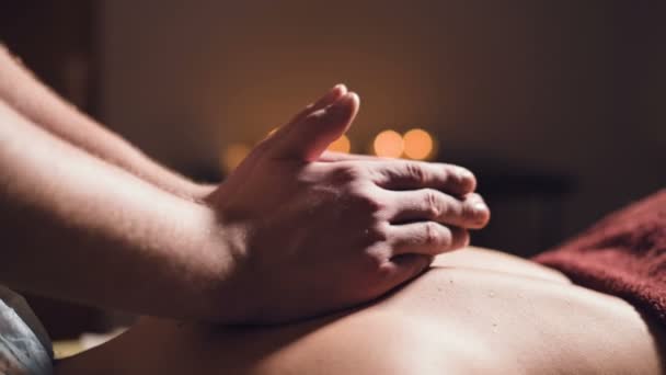 Close-up Jonge mannelijke massage therapeut doet rug massage aan een vrouw met een tatoeage in een massageruimte met schemerige lichten op de achtergrond van kaarsen. Low-key premium massageconcept — Stockvideo