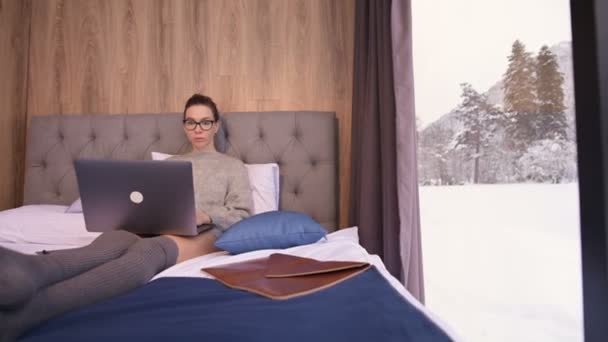 在一个有透明墙壁的生态旅馆里，一个戴着眼镜，床上有笔记本电脑的迷人的自由职业姑娘的画像，后面是一片冬季的森林。自由时间的旅行和工作概念. — 图库视频影像