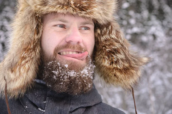 Πορτρέτο ενός γενειοφόρου άνδρα σε ένα γούνινο μεγάλο καπέλο στο φόντο ενός χιονισμένου δάσους. Χαμογελαστός άντρας στο δάσος. Δείχνει γλώσσα να γλείφει το μουστάκι του — Φωτογραφία Αρχείου
