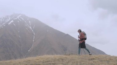 Sırt çantası ve takip çubukları olan genç bir adam bulutlu havada dağlarda alçak bulutların ve dağların arasında bir tepeye doğru yürüyor. Yürüyüş ve yürüyüş konsepti