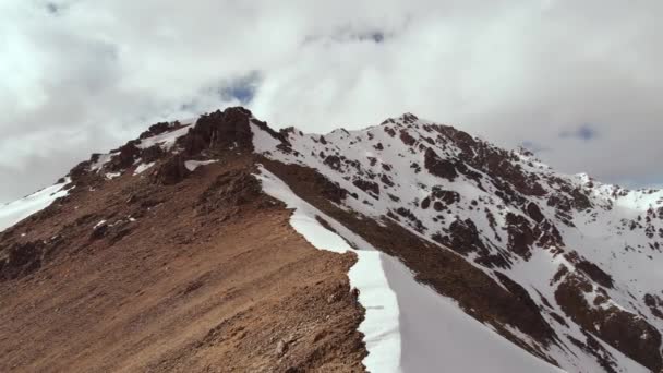 Vista aérea de un joven delgado con una mochila y bastones de trekking sube colina arriba en una cresta en el borde de rocas y rocas con nieve sobre el fondo de picos nevados y nubes — Vídeos de Stock