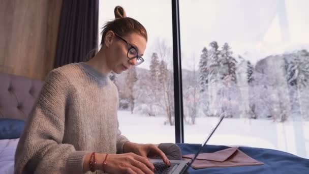 Une femme indépendante avec des lunettes est assise sur un lit dans une maison de campagne avec des fenêtres panoramiques derrière lesquelles il y a une forêt enneigée. Fille avec ordinateur portable ne fonctionne à distance tout en étant isolé — Video