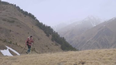 Sırt çantası ve takip çubukları olan genç bir adam bulutlu havada dağlarda alçak bulutların ve dağların arasında bir tepeye doğru yürüyor. Yürüyüş ve yürüyüş konsepti