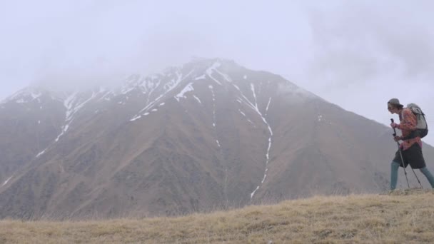Egy fiatal férfi hátizsákkal és nyomkövető botokkal felsétál egy dombra a hegyekben felhős időben, alacsony felhők és hegyek közepette. A kirándulás és a túrázás fogalma — Stock videók