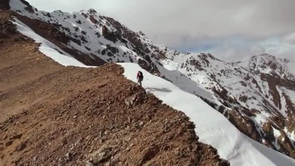 Flygfoto av en ung tunn man med en ryggsäck och vandringsstavar springa klättrar uppför ett krön på gränsen mellan stenar och stenar med snö mot bakgrund av snötäckta toppar och moln — Stockvideo