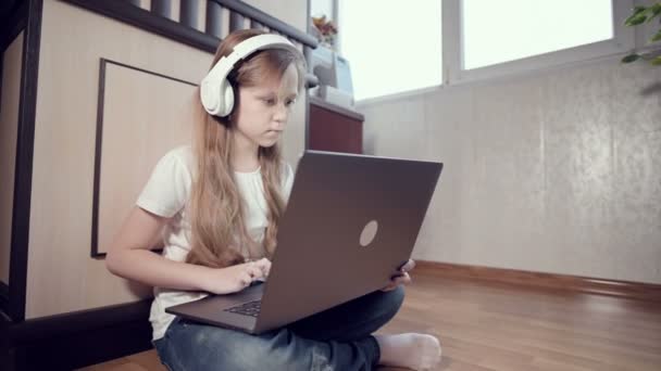 Una niña inteligente de siete años con auriculares blancos con un portátil en las manos está empujando al suelo en su habitación. La generación joven en Internet y la tecnología informática — Vídeo de stock