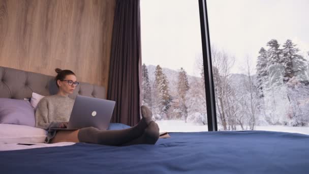 Portrét atraktivní dívky na volné noze v brýlích s laptopem na posteli v eko-hotelu s průhlednými stěnami, za nimiž je zimní les. Cestovní a pracovní koncept s volným termínem. — Stock video