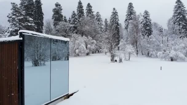 Vista aérea de una innovadora casa de alta tecnología con paredes transparentes espejadas en invierno cerca de un bosque de coníferas en las montañas del Cáucaso o los Alpes. — Vídeos de Stock