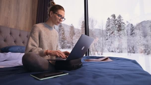 Portrait d'une jolie pigiste dans des lunettes avec un ordinateur portable sur le lit dans un éco-hôtel avec des murs transparents derrière lesquels il y a une forêt d'hiver. Concept de voyage et de travail avec horaire gratuit. — Video