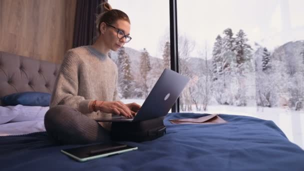 Retrato de atractiva chica freelancer en gafas con un portátil en la cama en un eco-hotel con paredes transparentes detrás de las cuales hay un bosque de invierno. Concepto de viaje y trabajo con horario libre. — Vídeos de Stock