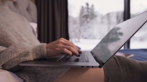 Zbliżenie dziewczyny ręce wydrukować coś na laptopie siedzi na łóżku z laptopem w rękach przed oknami panoramicznymi, za którymi zimowy las w śniegu. — Wideo stockowe