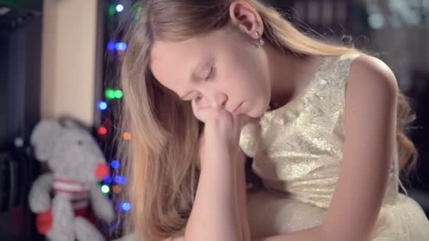 お祝いの光とクリスマスツリーを背景にお祝いのドレスの女の子は悲しいことに焦点を当てるためにぼかしや動きの下どこかを探します。甘やかされた休日の概念 — ストック動画