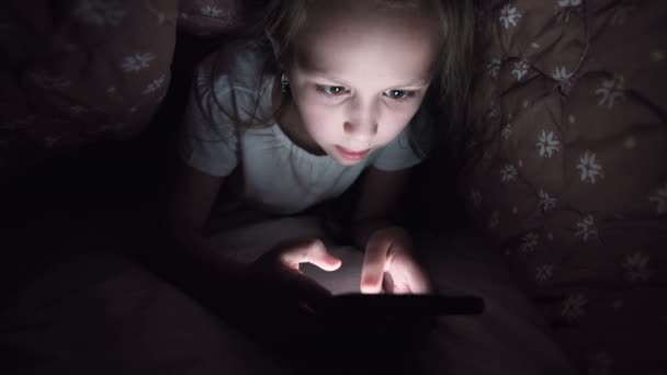Остроумная маленькая девочка играет на смартфоне ночью под одеялом. Чайлдс уходит от реальности через мобильные устройства. Ребенок пользуется секретным телефоном — стоковое видео