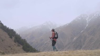 Sırt çantası ve takip çubukları olan genç bir adam bulutlu havada dağlarda alçak bulutların ve dağların arasında bir tepeye doğru yürüyor. Yürüyüş ve yürüyüş konsepti