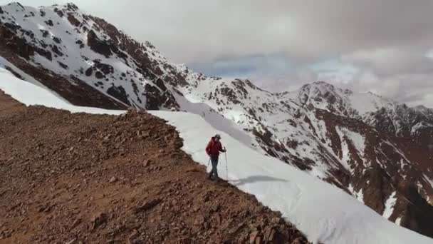 Luftbild junger Mann mit Sonnenbrille und Rucksack geht den Berg hinunter auf den Kamm an der Grenze zwischen Schnee und Steinen vor der Kulisse schneebedeckter Berge. — Stockvideo