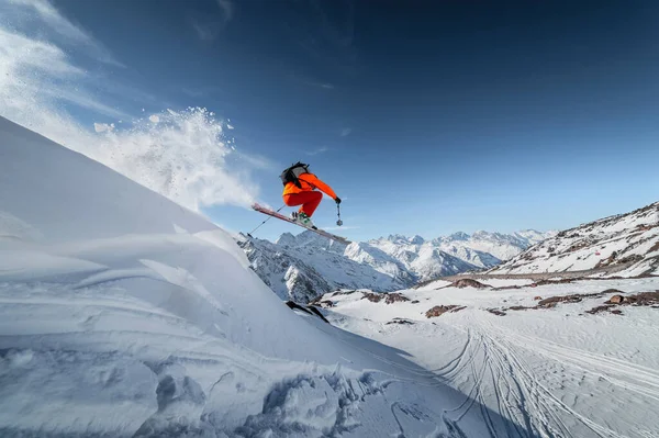 Athlete male skier jumps from a snow-covered slope against the backdrop of a mountain landscape of snow-covered mountains on a sunny day. The concept of winter sports wide angle — Stock Photo, Image