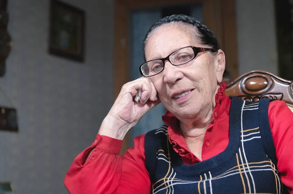 Retrato de una vieja mujer bien arreglada con gafas se sienta y sonríe slyly en una silla en la habitación —  Fotos de Stock