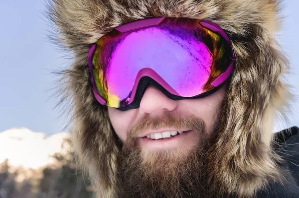 Ritratto ravvicinato di uno sciatore di snowboarder felice barbuto in un passamontagna con occhiali e una pelliccia grande cappello old-school su uno sfondo di montagne innevate invernali — Foto Stock