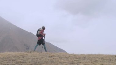 Sırt çantası ve takip çubukları olan genç bir adam bulutlu havada dağlarda alçak bulutların ve dağların arasında bir tepeye doğru yürüyor. Yürüyüş ve yürüyüş konsepti