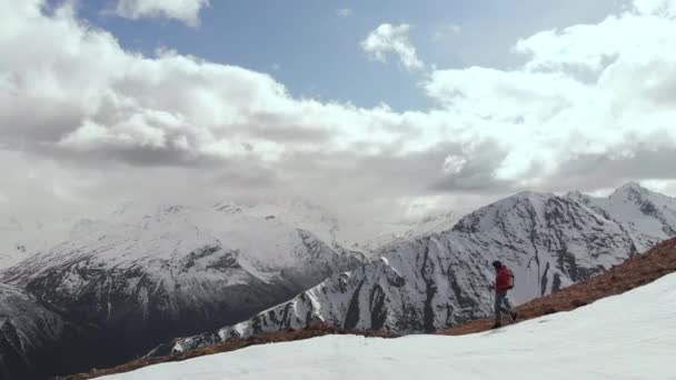Vista aérea jovem em óculos de sol com uma mochila desce a montanha na crista na borda da neve e pedras contra o pano de fundo de montanhas cobertas de neve. — Vídeo de Stock