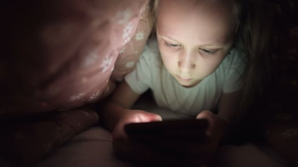 En pigg liten flicka spelar på en smartphone på natten under en filt. Barnen lämnar verkligheten via mobila enheter. Barn använder hemlig telefon — Stockvideo