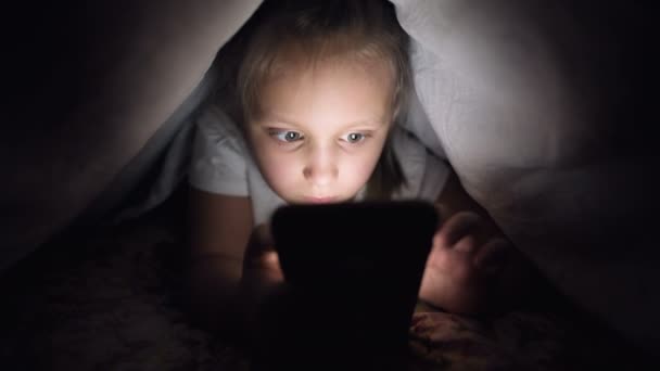 Una bambina appassionata gioca su uno smartphone di notte sotto una coperta. I bambini partono dalla realtà attraverso i dispositivi mobili. Bambino utilizza il telefono segreto — Video Stock