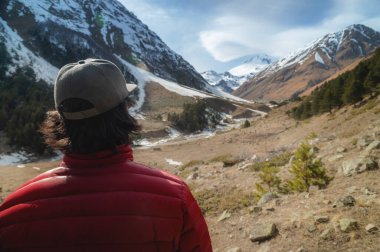 Uzun saçlı, kırmızı ceketli ve güneş gözlüklü gri şapkalı genç adam sırtı kameraya dönük duruyor ve uzaktaki karlı zirveleri olan dağ geçidine bakıyor.