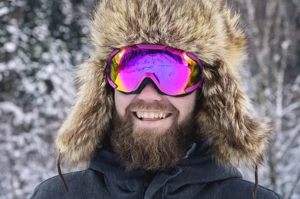 Retrato de cerca de un esquiador barbudo feliz snowboarder en una máscara de esquí con gafas y un sombrero de piel grande de la vieja escuela sobre un fondo de montañas nevadas de invierno —  Fotos de Stock