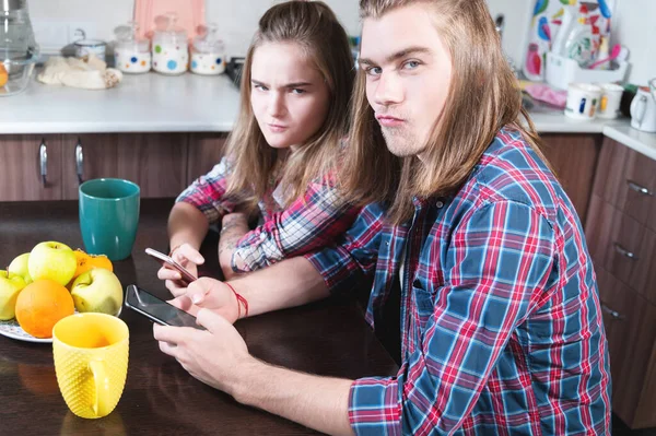 A young smile couple guy and a girl with long hair sit at the dinner table in the kitchen and use phones communicating with someone in chat rooms and social networks. — Stock Photo, Image