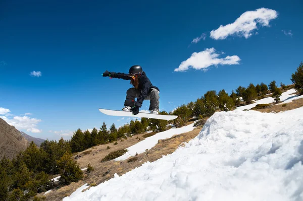 Стильная молодая девушка-сноубордистка делает трюк, прыгая со снегохода на фоне голубого неба облаков и гор весной. — стоковое фото