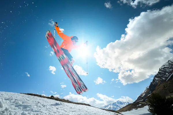 Een jonge stijlvolle man met een zonnebril en een pet doet een truc om op een zonnige dag met een sneeuwkick tegen de blauwe lucht en de zon te springen. Het concept van parkskiën in de winter of het voorjaar — Stockfoto