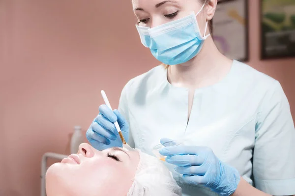 Μια αισθητικοί γραφείο μια γυναίκα αισθητικός βάζει μια βούρτσα αναζωογονητική μάσκα στο πρόσωπο ενός ελκυστικού πελάτη. Επαγγελματικές καλλυντικές υπηρεσίες — Φωτογραφία Αρχείου