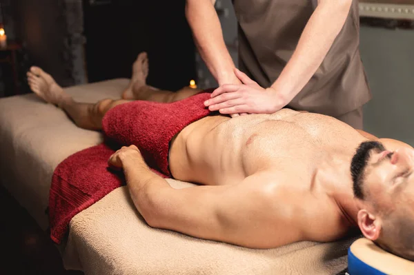 Male masseur massages the thoracic diaphragm of a muscular male athlete in a massage room with dark lighting